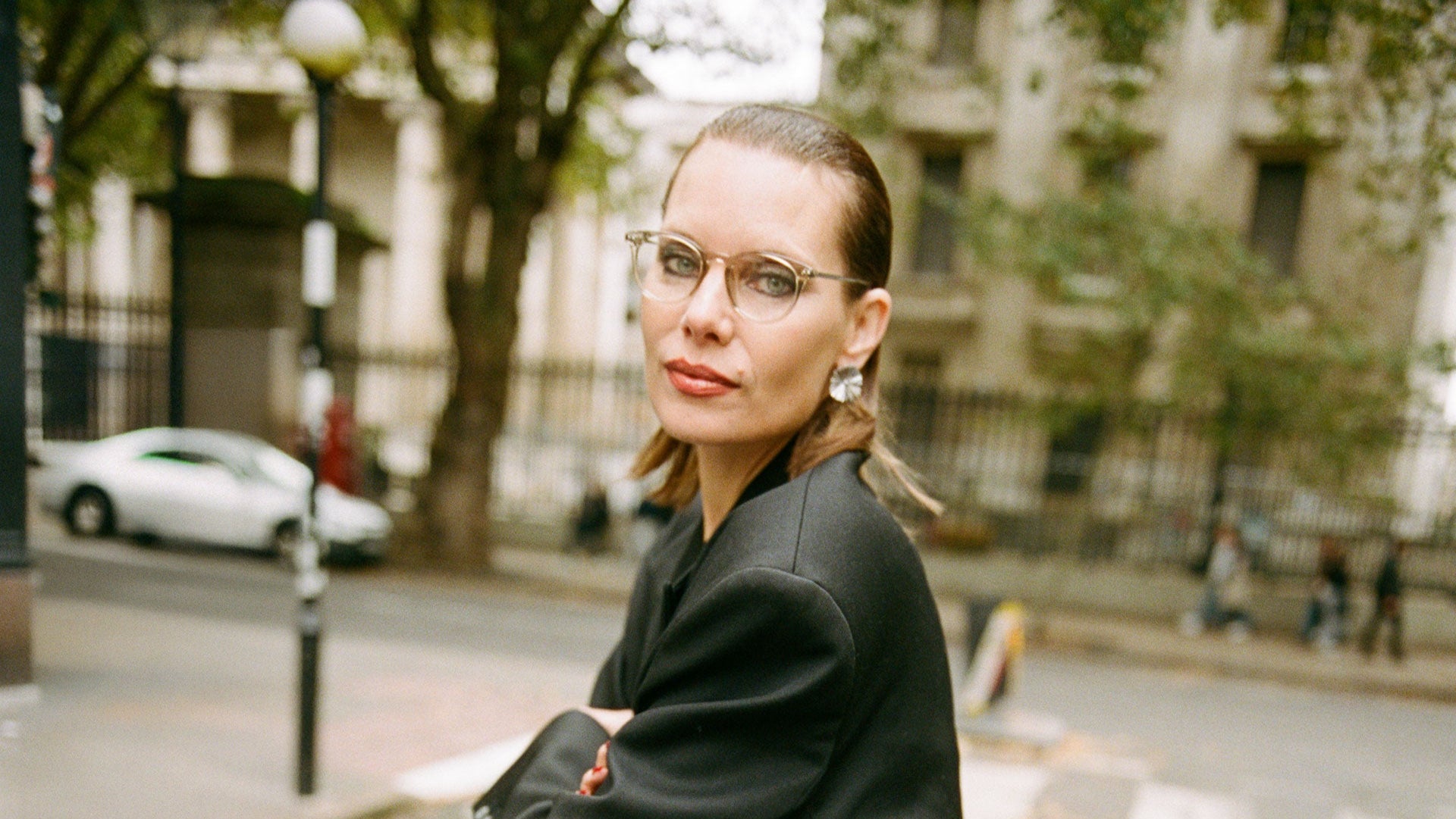 Woman in a black blazer and white pants standing on a street with trees and buildings in the background.